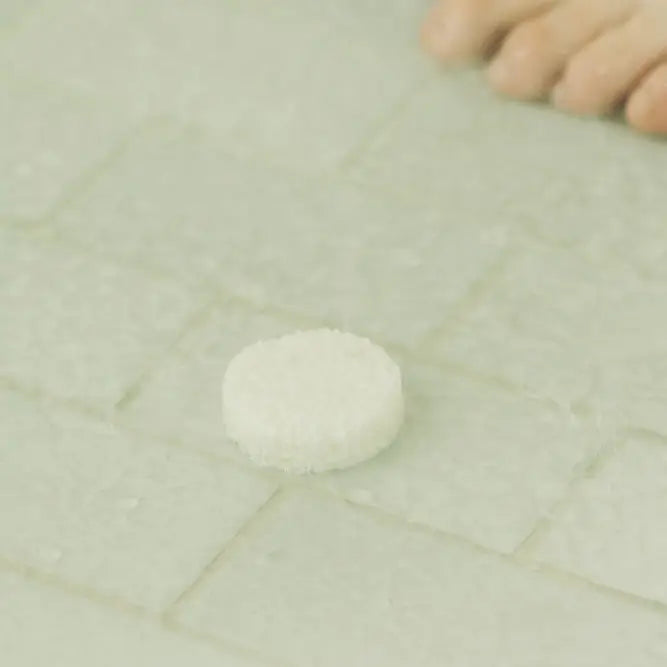 A small round white textured item placed on a pale tiled surface, with a blurred human foot visible in the upper right corner.