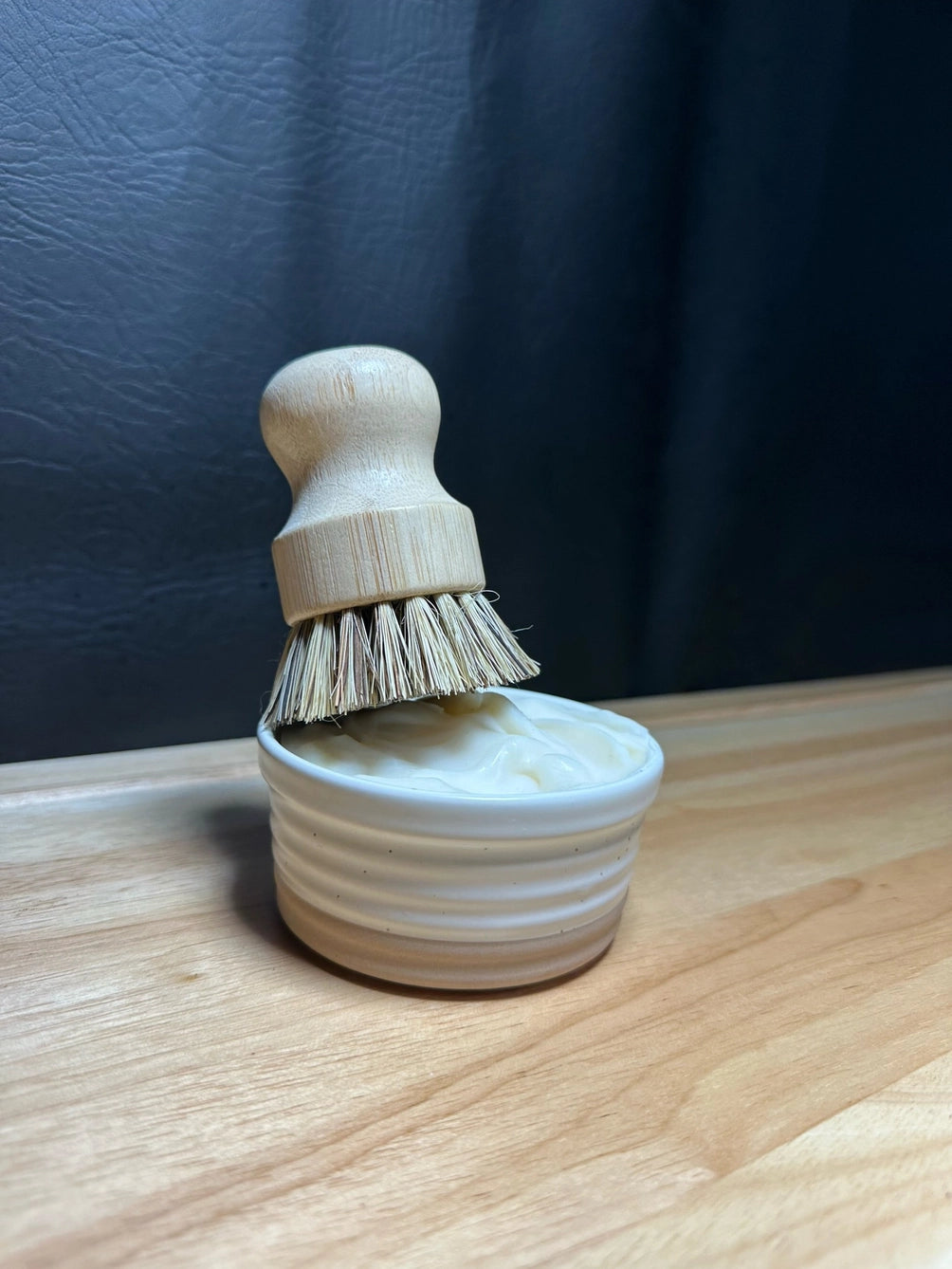A small wooden shoe brush with natural bristles resting in a ribbed cream-colored wooden cup, set on a light wood surface against a dark textured background.