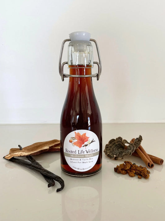 A glass swing-top bottle filled with amber liquid, labeled 'Rooted Life Wellness' and 'Cinnamon & Cloves', displayed on a light surface with cinnamon sticks, cloves, and vanilla beans around it.