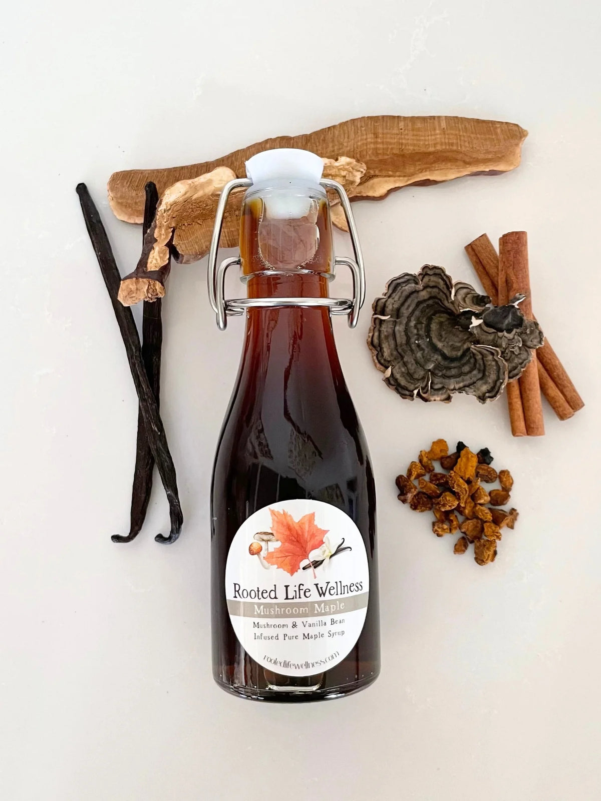 A glass swing-top bottle of Rooted Life Wellness Isoflavone & Vanilla Bean Infused Maple Syrup, styled with vanilla beans, cinnamon sticks, a mushroom, and dried botanicals on a light background.