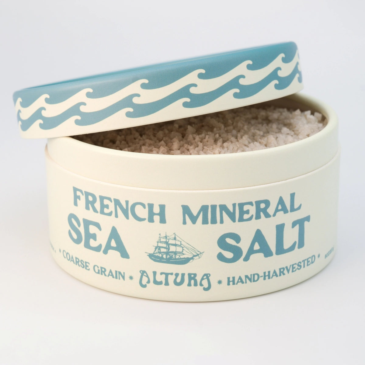 A round cream-colored tin of Altura French Mineral Sea Salt, shown open with its lid propped up. The lid features a blue and white wave pattern, and the tin is printed with 'FRENCH MINERAL SEA SALT' and a small sailing ship illustration in blue.