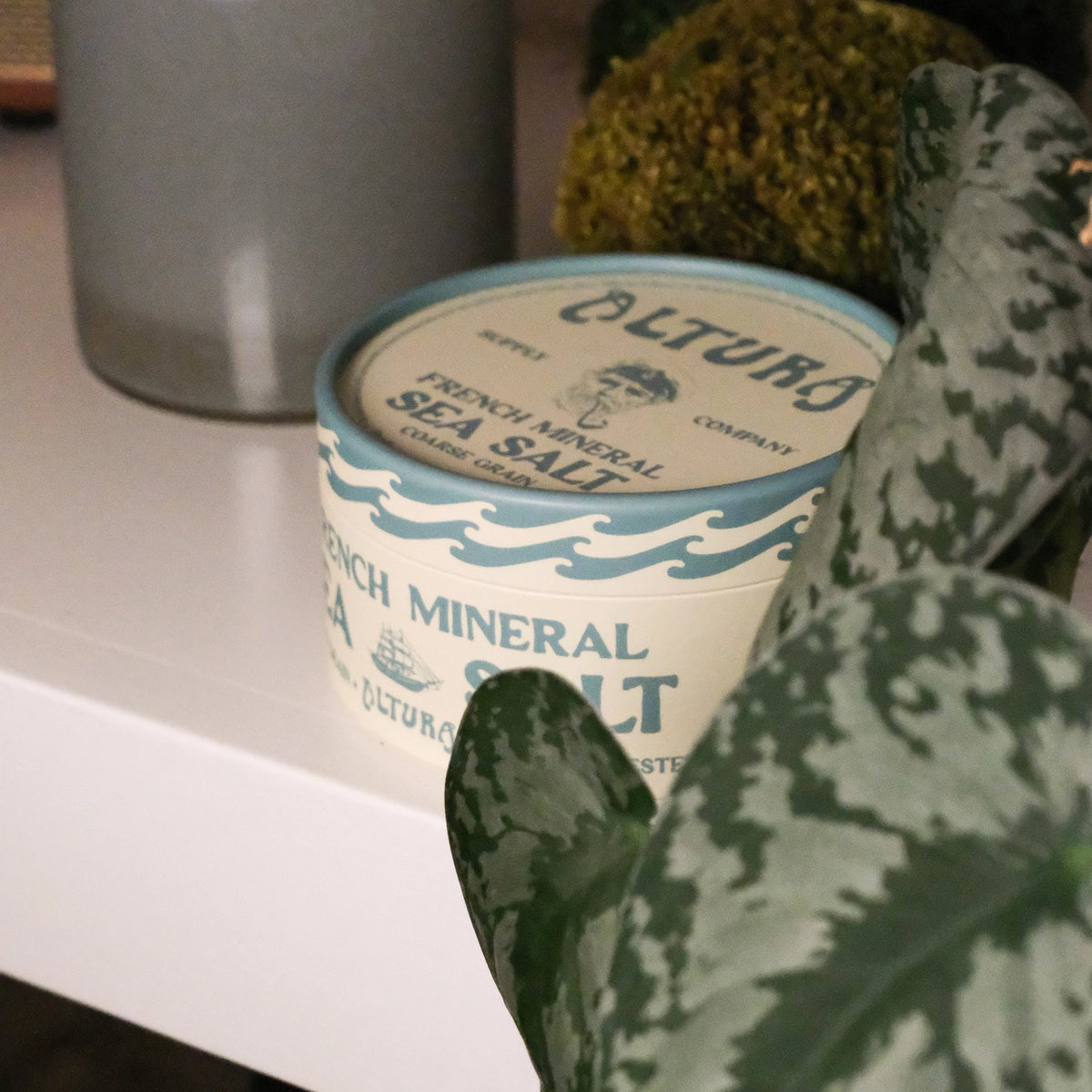 A close-up of a white plastic tub of Altura French Mineral Sea Salt, partially framed by a green patterned plant leaf in the foreground, set on a white shelf.
