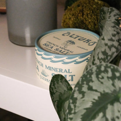 A close-up of a white plastic tub of Altura French Mineral Sea Salt, partially framed by a green patterned plant leaf in the foreground, set on a white shelf.