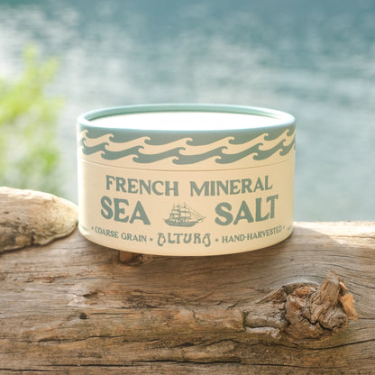A round tin of French Mineral Sea Salt sits on a weathered wooden log with a blurred body of water and green foliage in the background. The tin has a cream-colored label with dark green text and wave designs.