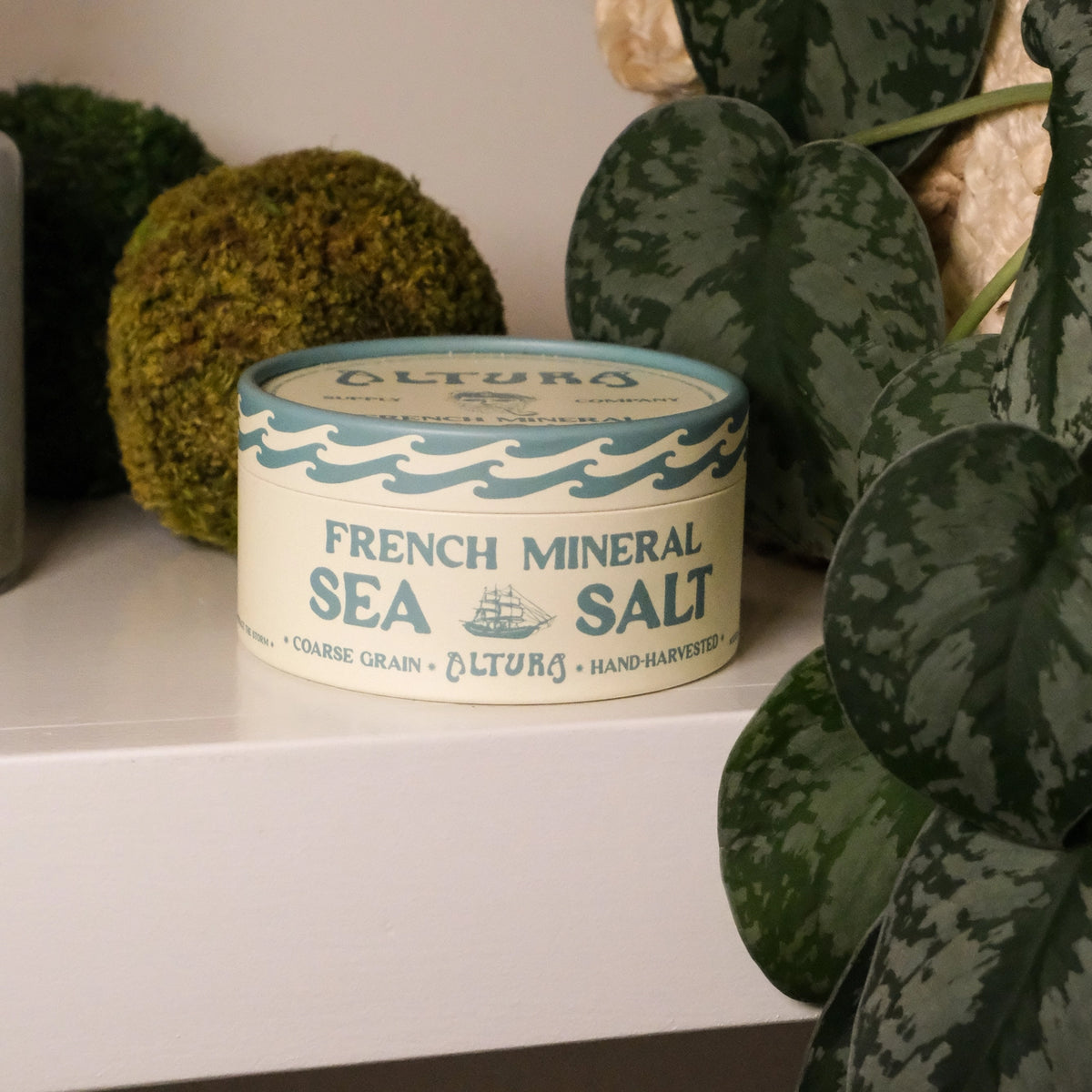 A tin of Altura French Mineral Sea Salt sits on a white shelf, styled with green moss balls and a houseplant in the background.