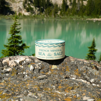 A can of Ocean Salt French Mineral Sea Salt sits on a rocky foreground with a turquoise lake and pine trees in the background.