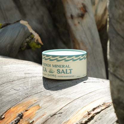 A small round tin of French Mineral Sea Salt sits on a weathered log outdoors, with blurred tree trunks in the background. The tin is cream-colored with a teal wave pattern and dark teal text.