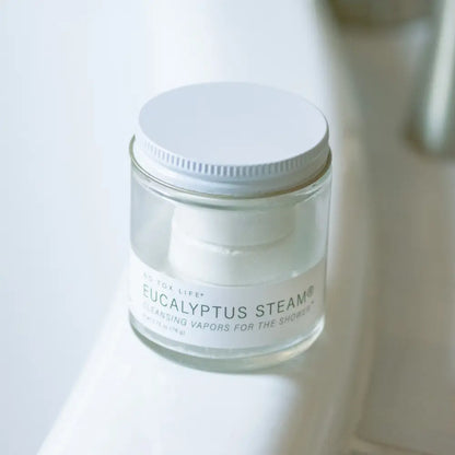 A clear glass jar of Eucalyptus Steam cleansing vapor with a white screw-on lid, photographed on a white surface with a softly blurred bathroom background.