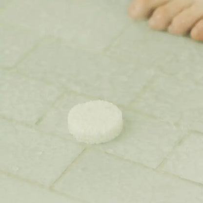 A small round white textured item placed on a pale tiled surface, with a blurred human foot visible in the upper right corner.