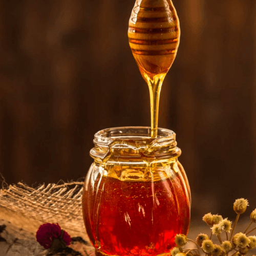 A honey dipper/wand dripping raw organic honey into a small glass jar, one of our most important ingredients in making the best organic elderberry syrup recipe to boost the immune system.