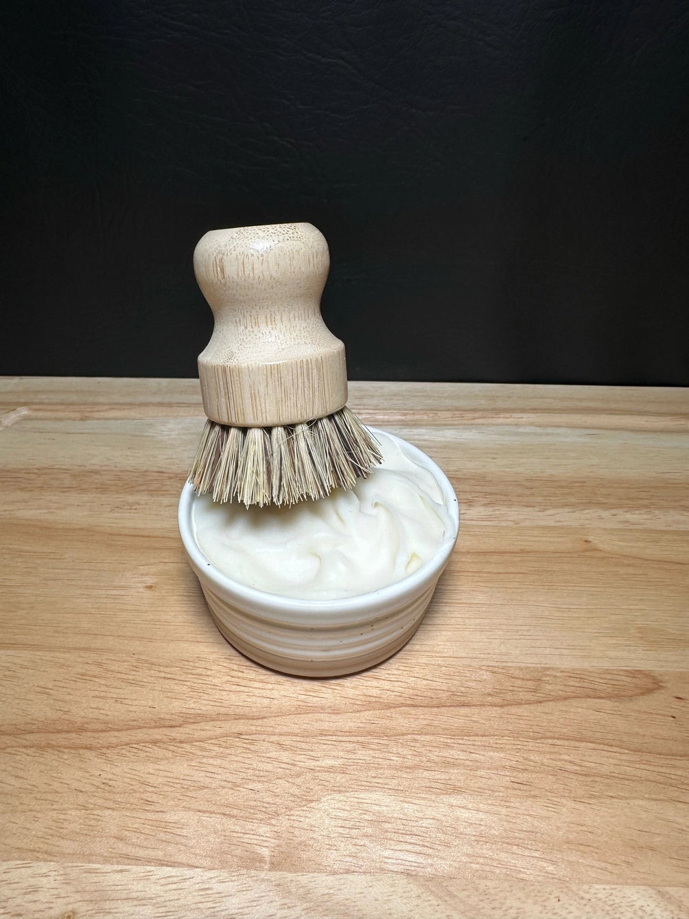 A natural wood grooming brush with soft bristles resting in a small white ceramic bowl filled with tallow dish soap, set on a light wood surface against a dark background.