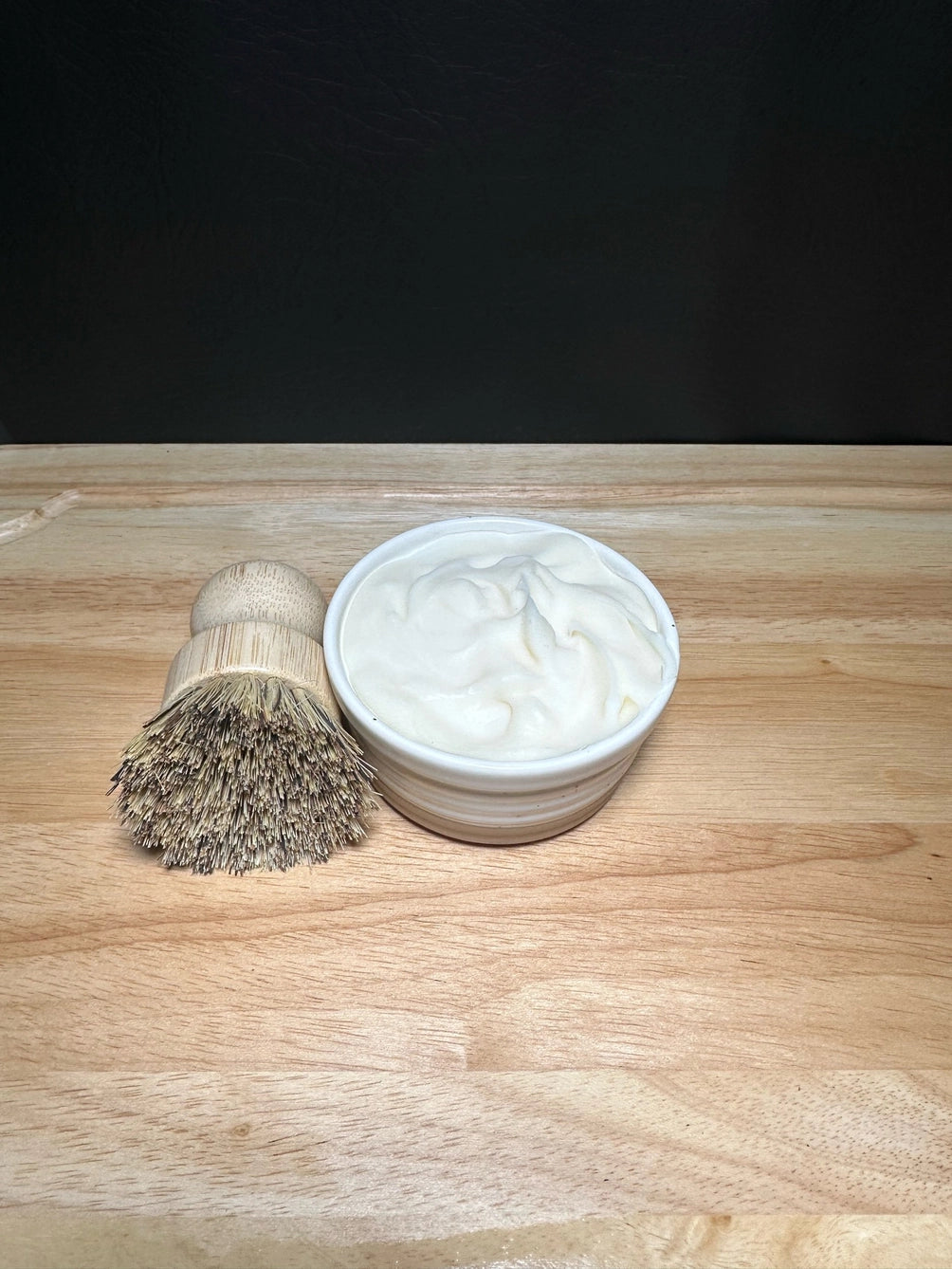 A small white ceramic bowl filled with tallow dish soap sits on a light wood surface next to a shaving brush with a natural wood handle and dark bristles, against a dark background.