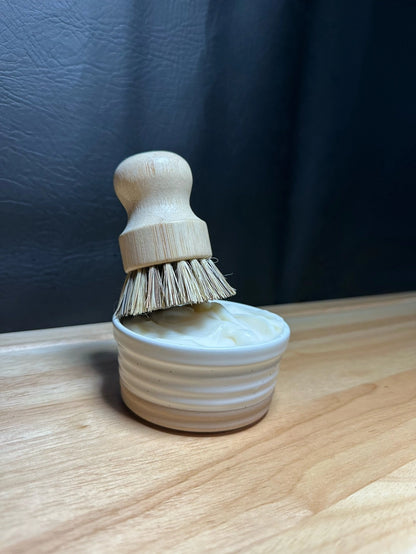 A small wooden shoe brush with natural bristles resting in a ribbed cream-colored wooden cup, set on a light wood surface against a dark textured background.