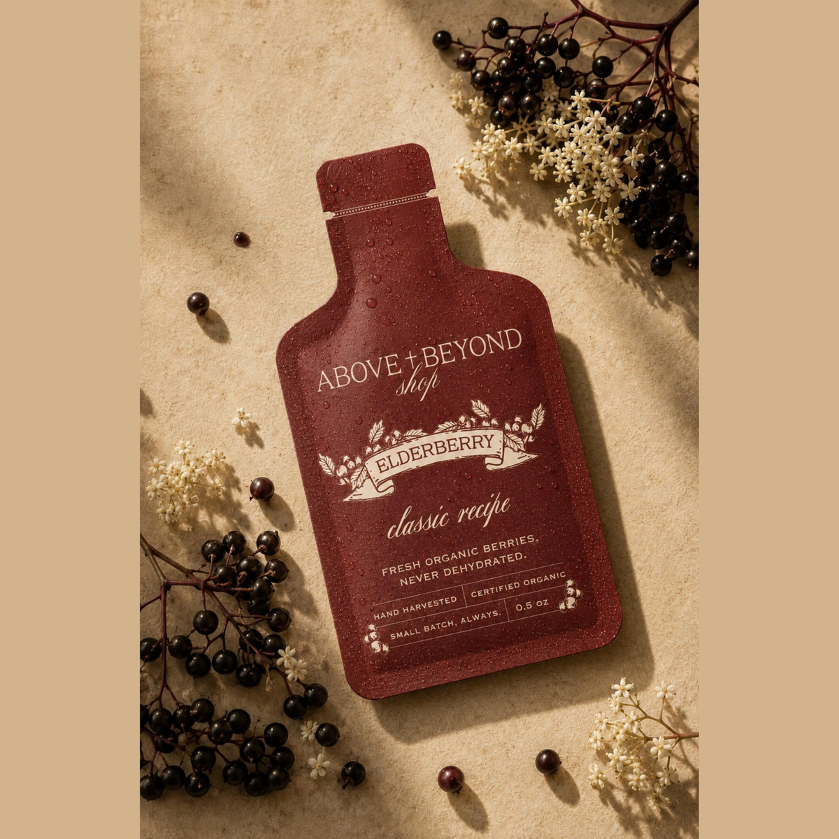 A single bottle-shaped elderberry supplement packet from Above + Beyond Shop, in a deep burgundy color with cream text, styled on a textured beige background with dried elderberries and white flowers.