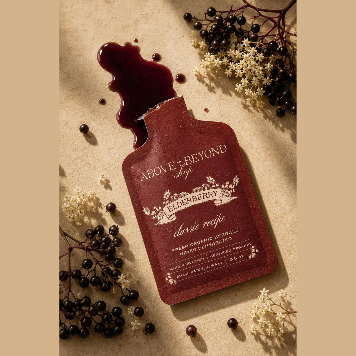 A single elderberry pouch package lies on a beige surface with dried elderberry berries and white flowers arranged around it. The pouch is dark red/burgundy with white text reading 'ABOVE + BEYOND shop ELDERBERRY classic recipe.