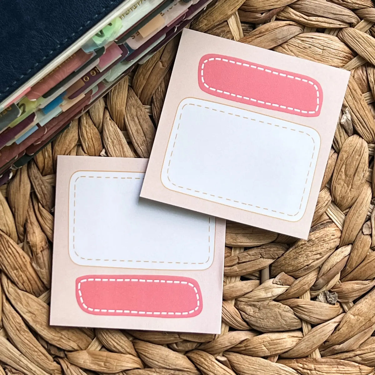 Two small beige sticky note pads with pink rounded label areas and dashed border details, arranged on a woven straw surface with a stack of cards partially visible at the top left.