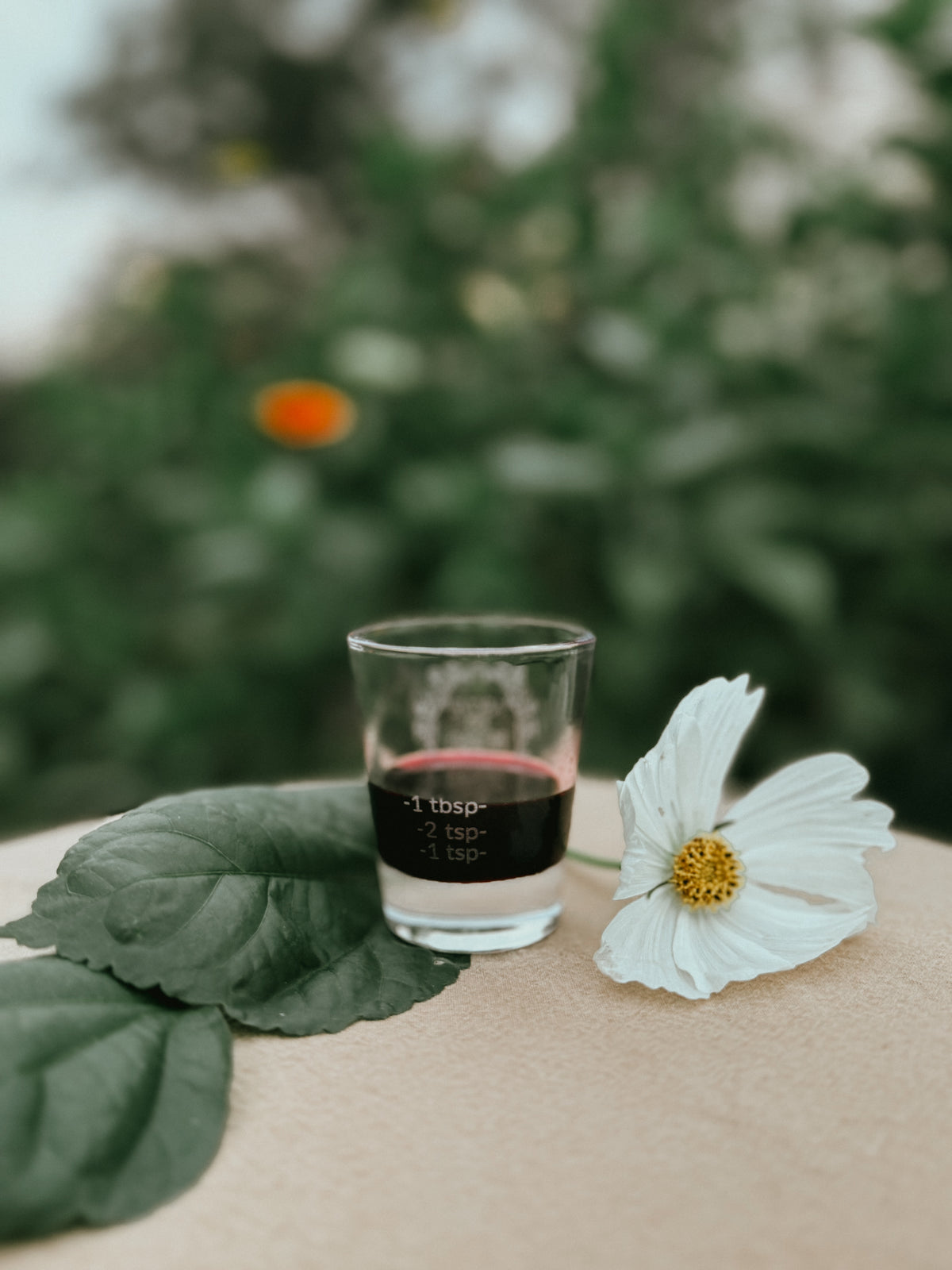 A small clear glass shot glass with a black printed measurement label, placed on a beige surface beside a white flower and green leaves, with a blurred green outdoor background.