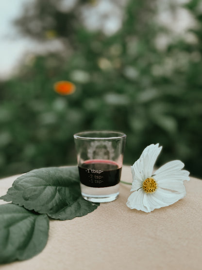 A small clear glass shot glass with a black printed measurement label, placed on a beige surface beside a white flower and green leaves, with a blurred green outdoor background.