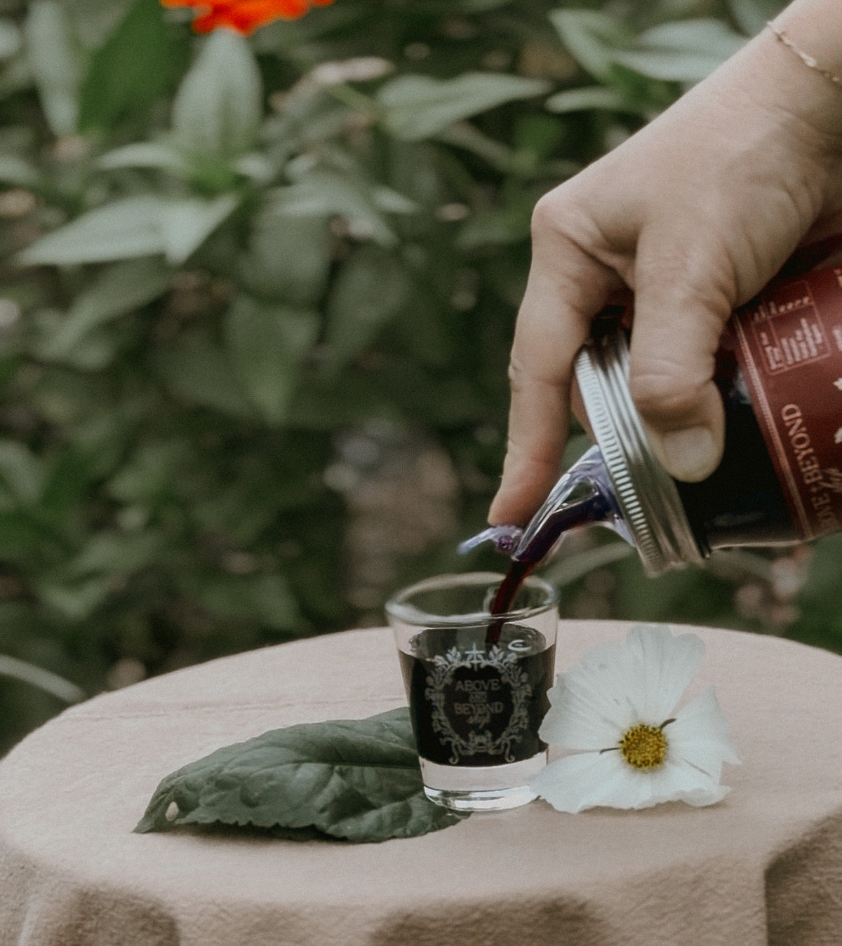 A hand pours a dark red liquid from a bottle into a small clear glass on a beige tablecloth, with a white flower and green leaves in the foreground and blurred green foliage in the background.