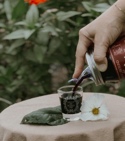 A hand pours a dark red liquid from a bottle into a small clear glass on a beige tablecloth, with a white flower and green leaves in the foreground and blurred green foliage in the background.