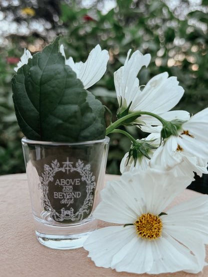 A clear glass tumbler with white floral engraving reading 'ABOVE AND BEYOND shop' sits on a beige surface outdoors, holding a green leaf and white cosmos flowers against a blurred green garden background.