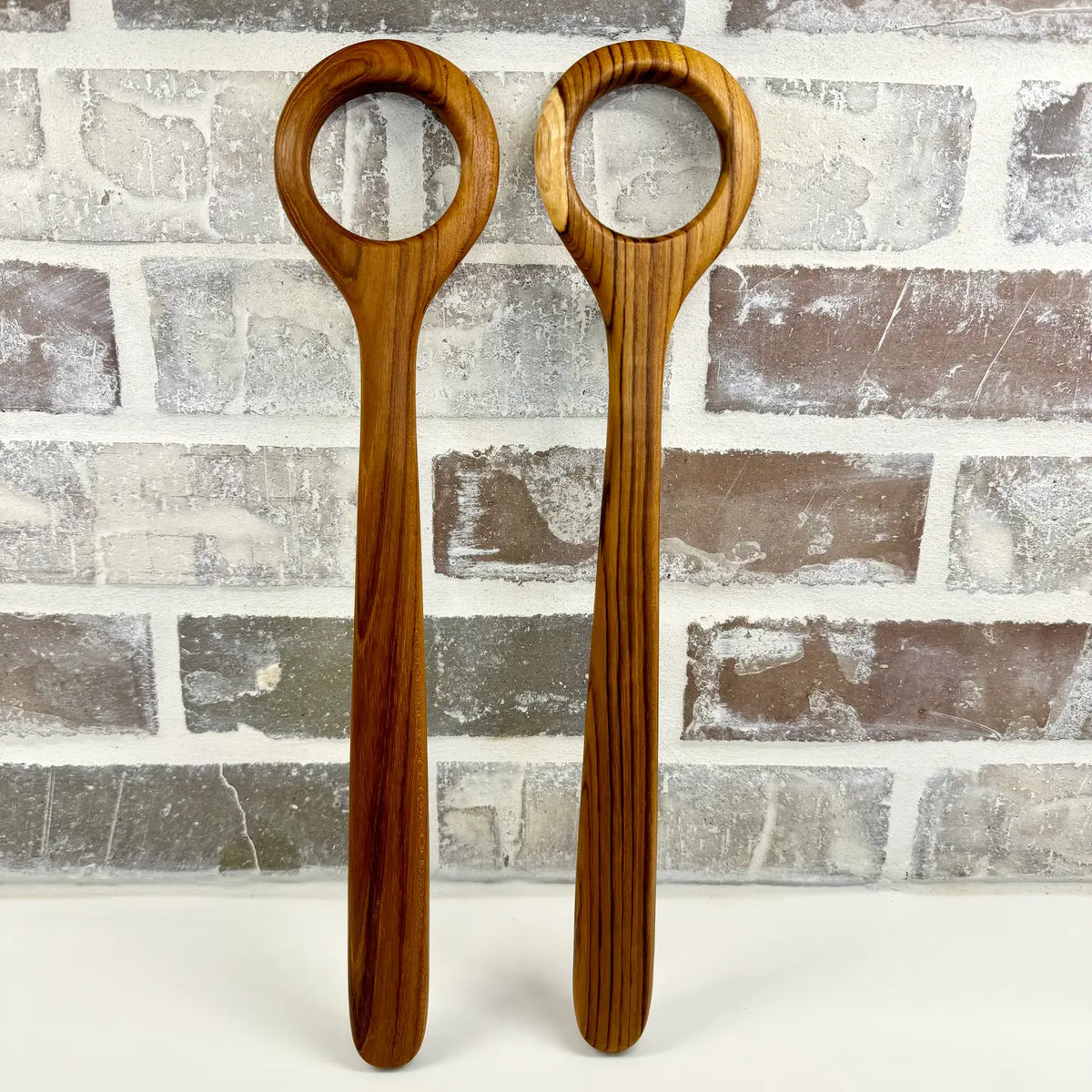 Two wooden dough whisks with circular bowls, standing upright against a faux brick wall background.