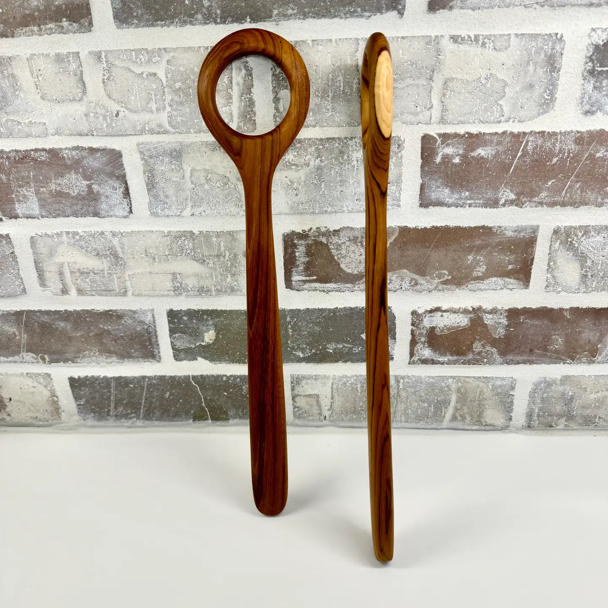 Two wooden dough whisks with a distinctive loop handle and oval spoon bowl, standing upright against a gray brick wall background.