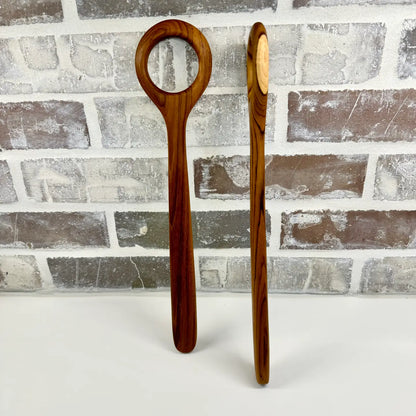 Two wooden dough whisks with a distinctive loop handle and oval spoon bowl, standing upright against a gray brick wall background.