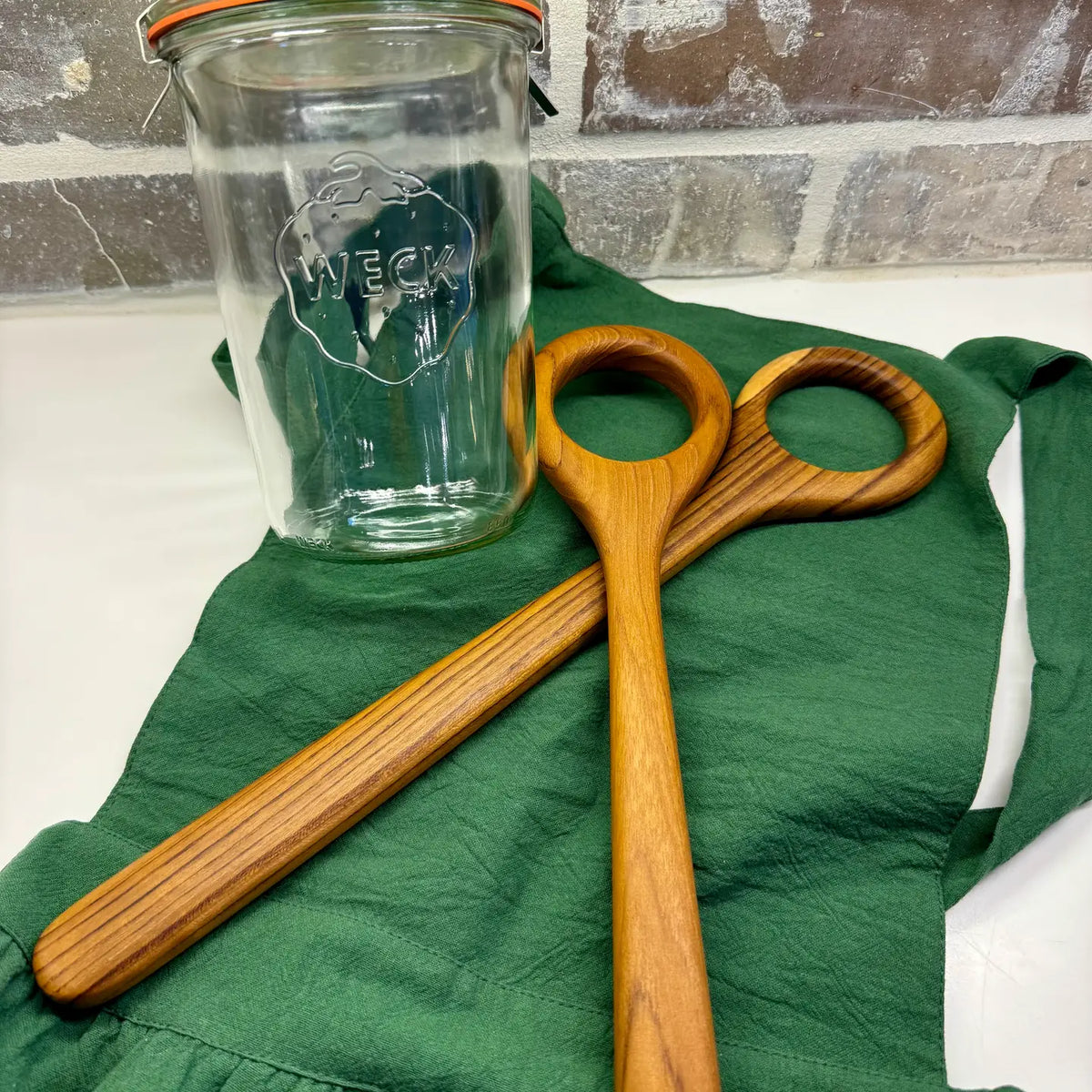 A clear glass jar with a metal lid and a visible WECK logo, placed on a green cloth next to two wooden dough whisks with circular loops, against a brick-patterned background.