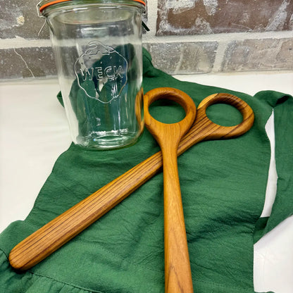 A clear glass jar with a metal lid and a visible WECK logo, placed on a green cloth next to two wooden dough whisks with circular loops, against a brick-patterned background.